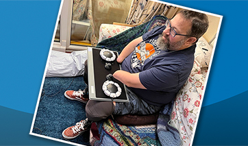 Seated man with beard operating two circular accessible games controllers on a laptray with both part-amputated arms