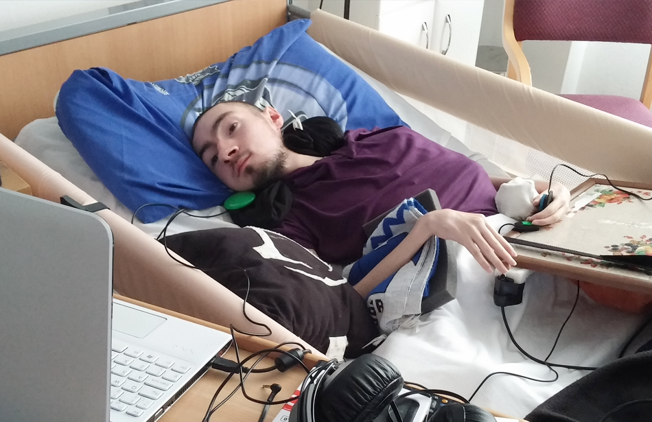 Young man lying in bed looking at a computer screen which he is controlling with chin and finger switches